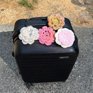 May include: A black suitcase with five colorful, crocheted flower appliques. The flowers are in shades of pink, white, yellow, and blue. The suitcase is sitting on a concrete surface.