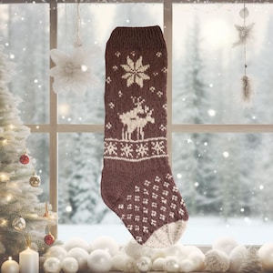 Peut inclure: Une chaussette de Noël marron avec un motif de rennes et de flocons de neige crème. Le bas tricoté présente un motif texturé et un bout crème. L'arrière-plan montre une scène hivernale enneigée.