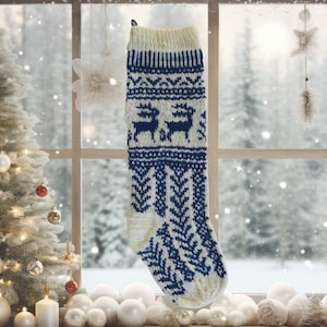 Pode incluir: Uma meia de Natal de malha em creme e azul marinho. A meia apresenta um padrão de renas e desenhos geométricos. A parte superior e o calcanhar são creme, enquanto o corpo tem um padrão azul marinho. Está pendurada em frente a uma janela.