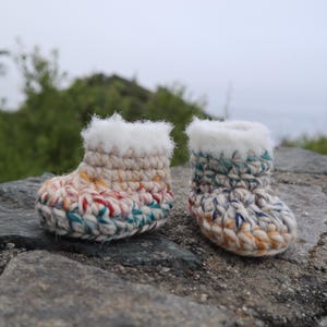 May include: Two handmade baby booties. The booties are crocheted with a colorful yarn in shades of cream, red, teal, and yellow. Each bootie has a fluffy white trim around the top. They are sitting on a gray stone surface.
