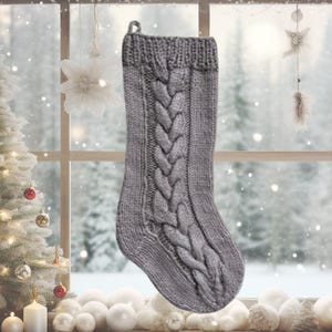 Può includere: Una calza di Natale grigia lavorata a maglia con un motivo a trecce. La calza ha un anello per appenderla ed è esposta davanti a una finestra con una scena invernale. È una decorazione festiva.