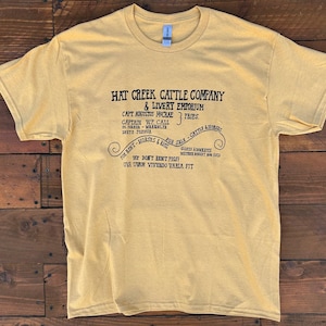 May include: Mustard yellow t-shirt with a crew neck and short sleeves. The front features black text that reads "Hat Creek Cattle Company & Livery Emporium" with additional text and decorative flourishes. The shirt is made of a soft material.