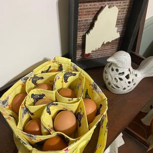 May include: A yellow fabric egg basket with a repeating rooster and hen print, featuring multiple interior compartments filled with brown chicken eggs. The basket has two fabric handles and a soft, structured design for holding eggs.