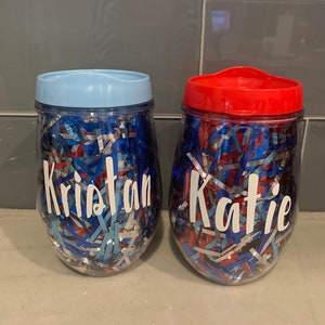 May include: Two clear plastic tumblers with red and blue confetti inside. The tumblers have lids, one blue and one red. The tumblers are personalized with the names "Kristan" and "Katie" in white vinyl lettering.