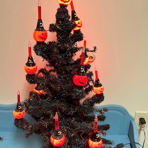 Può includere: Un albero di Halloween nero decorato con luci a forma di zucca arancione e lampadine rosse a forma di candela. L'albero è su una superficie blu, creando una decorazione festosa e spettrale.