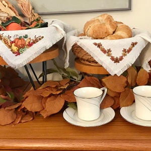 Peut inclure: Deux supports en bois à deux niveaux avec des serviettes en lin blanc brodées de motifs d'automne. Le niveau supérieur contient un panier de croissants et le niveau inférieur contient un panier d'articles de la récolte d'automne. Deux tasses à thé blanches avec des bordures argentées et des soucoupes assorties sont sur la table devant les supports. La table est recouverte de feuilles d'automne brunes.