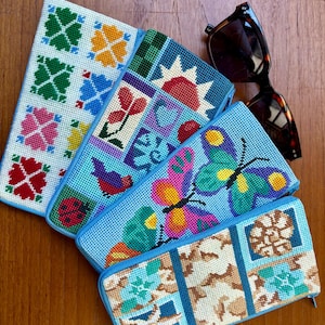 May include: Four rectangular zippered pouches featuring needlepoint designs. One has a white background with colorful floral motifs, another displays a sun and bird, a third shows a butterfly, and the last features seashells and starfish.