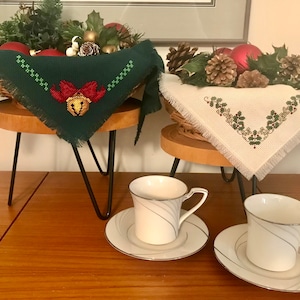 Puede incluir: Dos pequeños soportes de madera con patas de metal negro, cada uno con una cesta de mimbre con una tela bordada con motivos navideños. Las cestas están llenas de ramas de pino, piñas y adornos rojos. Dos tazas de té blancas con diseños azules y plateados están sobre la mesa delante de los soportes.