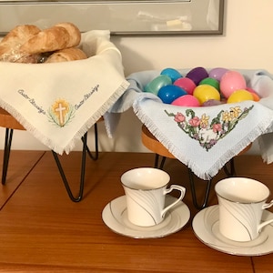 May include: Two white crocheted tablecloths with embroidered designs on black metal stands. One tablecloth has a cross and the words "Easter Blessings" embroidered on it. The other tablecloth has a bunny rabbit and flowers embroidered on it. There are colorful Easter eggs in the second tablecloth. Two white teacups with silver trim and matching saucers are on a wooden table.