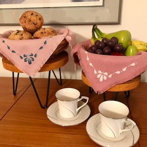 May include: Two wooden stands with black metal legs, each holding a basket with a pink cloth napkin. One basket contains muffins, the other contains fruit. Two white coffee cups with saucers are on the table.