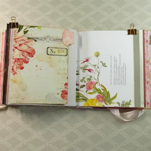 May include: An open scrapbook with two pages held by gold binder clips. The left page features a red floral illustration and a box labeled No. 8254. The right page displays botanical drawings of various flowers with text listing plant names.