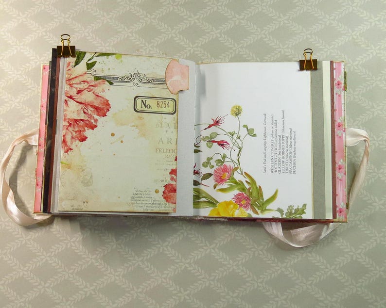 May include: An open scrapbook with two pages held by gold binder clips. The left page features a red floral illustration and a box labeled No. 8254. The right page displays botanical drawings of various flowers with text listing plant names.