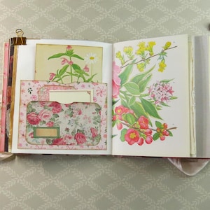 May include: An open scrapbook with pink ribbon ties, featuring a left page with a floral pocket containing a small card and a right page displaying a botanical illustration of yellow and red flowers. Gold metal clips secure the top edges of the pages.