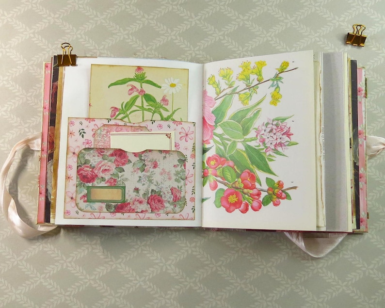 May include: An open scrapbook with pink ribbon ties, featuring a left page with a floral pocket containing a small card and a right page displaying a botanical illustration of yellow and red flowers. Gold metal clips secure the top edges of the pages.