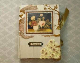 May include: A rectangular journal with a cream-colored fabric cover featuring a central rectangular print of a floral bouquet in a vase. The cover includes gold-toned botanical illustrations, a metal label plate reading Journal, and a cluster of cream-colored fabric flowers.