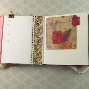 May include: An open scrapbook with white pages, a gold metal binder clip, and a decorative floral paper strip with lace trim. The right page features a vintage-style square card with a red rose illustration, cursive script, and a circular number 74.
