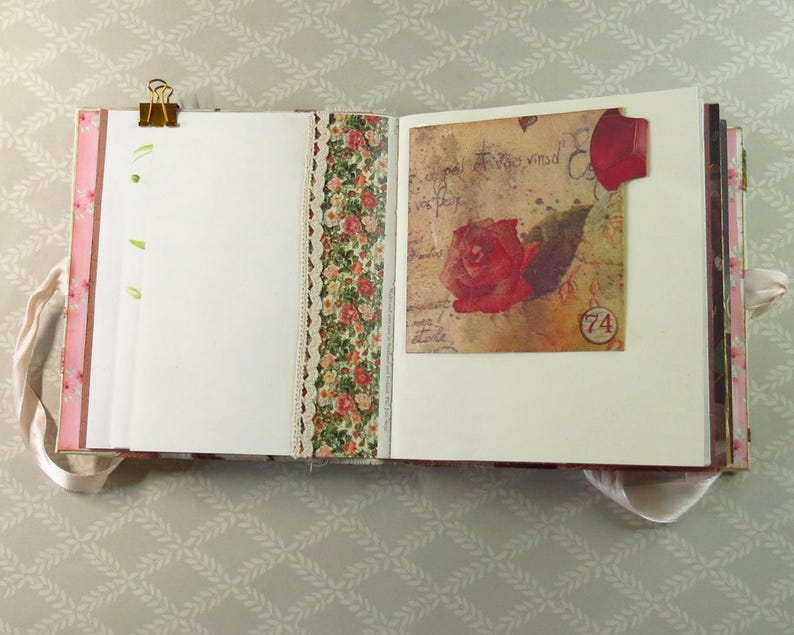 May include: An open scrapbook with white pages, a gold metal binder clip, and a decorative floral paper strip with lace trim. The right page features a vintage-style square card with a red rose illustration, cursive script, and a circular number 74.