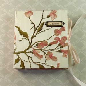 May include: A square journal with a cream-colored fabric cover featuring embroidered pink flowers and green leaves on brown branches. A rectangular metal label plate with the word Journal is attached to the top right. A light ribbon is tied to the side.