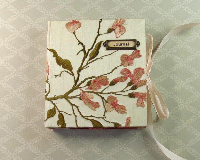 May include: A square journal with a cream-colored fabric cover featuring embroidered pink flowers and green leaves on brown branches. A rectangular metal label plate with the word Journal is attached to the top right. A light ribbon is tied to the side.