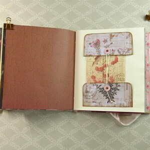 May include: An open scrapbook with a brown crackle-textured left page and a cream-colored right page featuring a central pocket system. The pockets are decorated with vintage floral and botanical illustrations, secured by a string wrapped around two pink buttons.