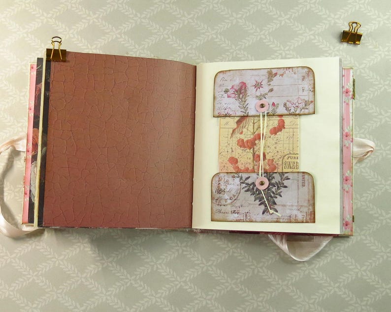May include: An open scrapbook with a brown crackle-textured left page and a cream-colored right page featuring a central pocket system. The pockets are decorated with vintage floral and botanical illustrations, secured by a string wrapped around two pink buttons.