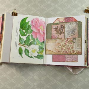 May include: An open scrapbook featuring a botanical illustration of pink and white camellia flowers on the left page and a layered collage of vintage-style paper tags, floral patterns, and handwritten script text on the right page.