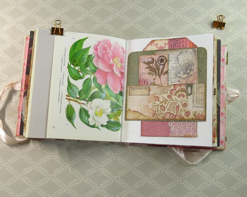 May include: An open scrapbook featuring a botanical illustration of pink and white camellia flowers on the left page and a layered collage of vintage-style paper tags, floral patterns, and handwritten script text on the right page.
