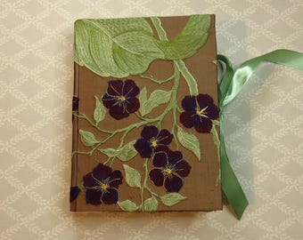 May include: A rectangular brown book with an embroidered design featuring large light green leaves and dark purple flowers with yellow centers. A light green satin ribbon is tied to the side of the book cover.
