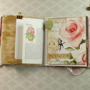 May include: An open scrapbook featuring a page with a vintage botanical illustration of a pink flower and printed text, alongside a page decorated with large watercolor-style pink roses and the numerical sequence 9876543210 in a rectangular box.