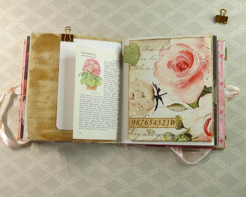May include: An open scrapbook featuring a page with a vintage botanical illustration of a pink flower and printed text, alongside a page decorated with large watercolor-style pink roses and the numerical sequence 9876543210 in a rectangular box.