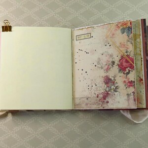 May include: An open journal with a gold metal binder clip on the left edge. The left page is blank and cream-colored. The right page features a vintage-style floral pattern with red roses, black ink splatters, and a small label reading OCT-12-23.