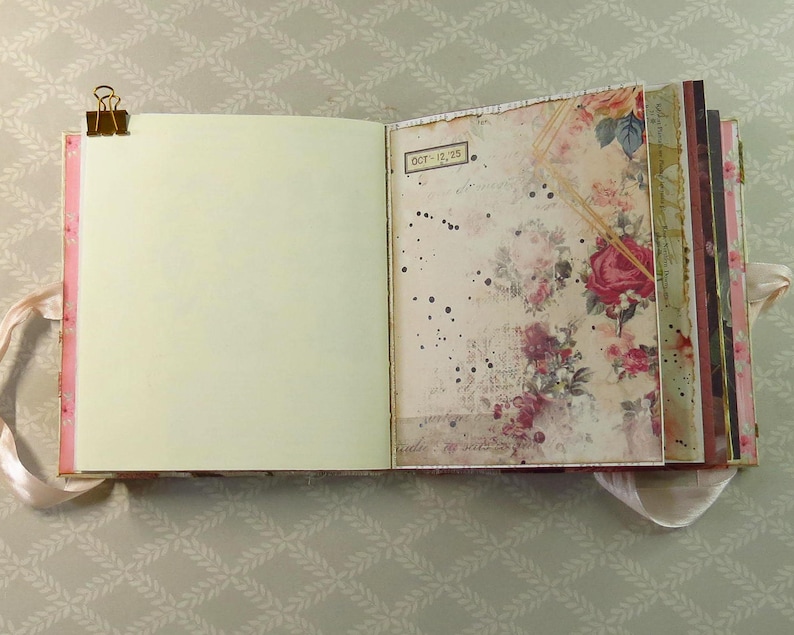 May include: An open journal with a gold metal binder clip on the left edge. The left page is blank and cream-colored. The right page features a vintage-style floral pattern with red roses, black ink splatters, and a small label reading OCT-12-23.