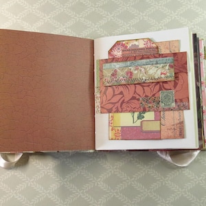 May include: An open scrapbook with a brown, crackled-texture left page and a right page featuring layered paper ephemera. The right side includes floral patterns, vintage-style script, a small green postage stamp, and a tag labeled Bains Douches.
