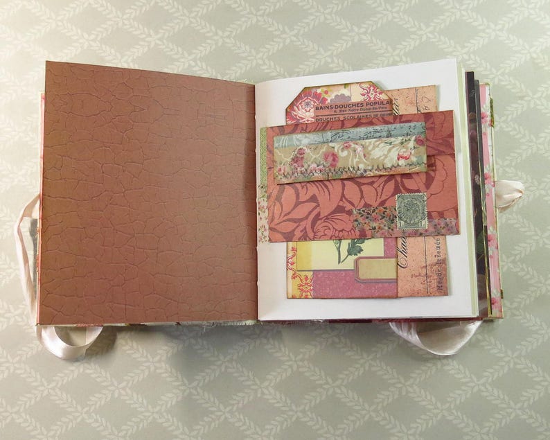 May include: An open scrapbook with a brown, crackled-texture left page and a right page featuring layered paper ephemera. The right side includes floral patterns, vintage-style script, a small green postage stamp, and a tag labeled Bains Douches.