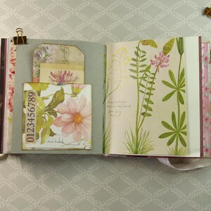 May include: An open scrapbook with a sage green pocket containing a floral tag and a strip of paper printed with the numbers 0123456789. The opposite page features a botanical illustration of green plants with pink and yellow flowers and handwritten text.