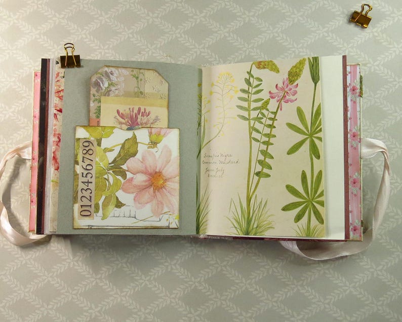 May include: An open scrapbook with a sage green pocket containing a floral tag and a strip of paper printed with the numbers 0123456789. The opposite page features a botanical illustration of green plants with pink and yellow flowers and handwritten text.