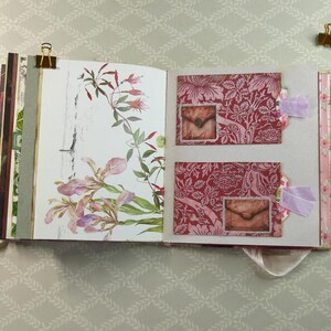 May include: An open scrapbook featuring a left page with a botanical illustration of pink and green flowers and a right page with two red patterned pockets, each containing a small envelope image, accented with light purple fabric tabs and gold clips.