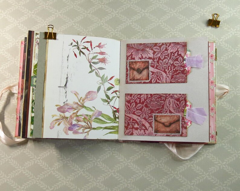 May include: An open scrapbook featuring a left page with a botanical illustration of pink and green flowers and a right page with two red patterned pockets, each containing a small envelope image, accented with light purple fabric tabs and gold clips.