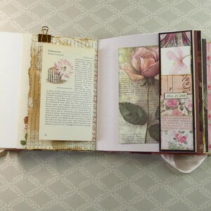 May include: An open scrapbook featuring pages with botanical illustrations, including a cactus with a pink flower and a large rose. The pages contain vintage-style text, handwritten script, and a small tag with the date July 17, 1906, held by a gold clip.