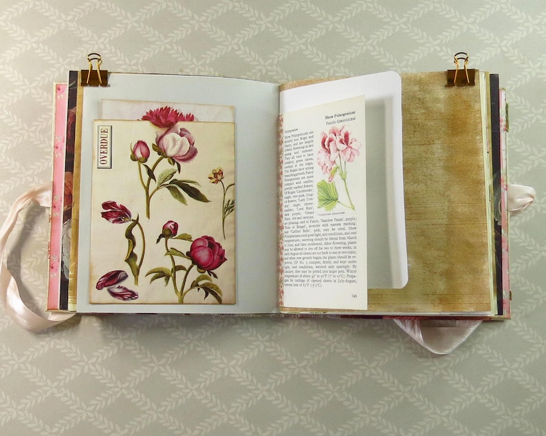 May include: An open scrapbook with two pages featuring vintage-style botanical illustrations of pink peonies and a printed page about Pelargonium plants. The pages are held open by gold metal binder clips and include a vertical pocket with the word OVERDUE.