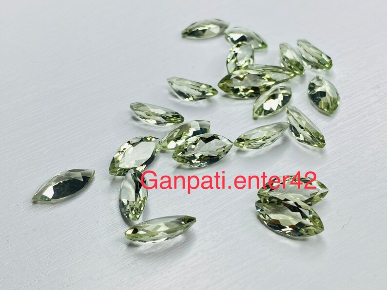 May include: A collection of light green marquise-shaped gemstones. The gemstones are scattered on a white surface.