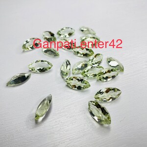 May include: A collection of light green marquise cut gemstones. The gemstones are scattered on a white surface.