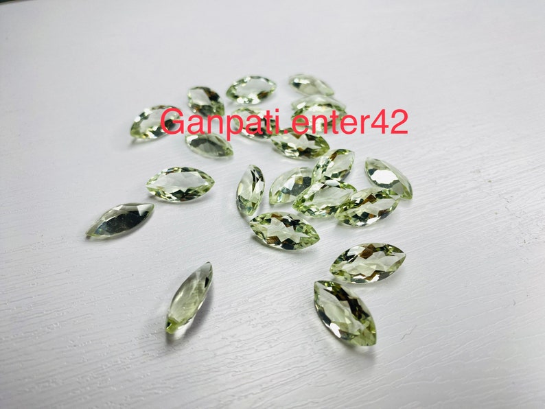 May include: A collection of light green marquise cut gemstones. The gemstones are scattered on a white surface.