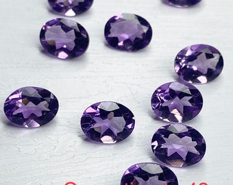 May include: Ten oval-shaped amethyst gemstones, each with a purple hue and a faceted cut. The gemstones are arranged in a scattered pattern on a white surface.