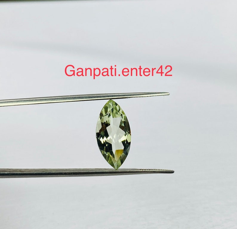 May include: A light green marquise cut gemstone held by tweezers. The gemstone is clear and has a slight yellow tint.