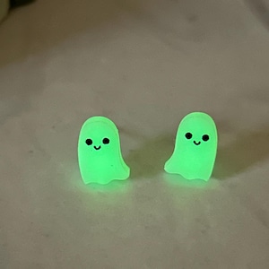 May include: Two glowing green ghost-shaped earrings with black eyes and smiling mouths. The earrings are small and have a smooth texture. The background is a light, neutral color.
