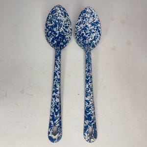 May include: Two enamel spoons with a blue and white speckled design. The spoons have long handles and oval bowls. The spoons are made of metal and have a hole at the end of the handle for hanging.