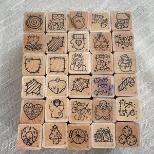 May include: A set of 25 wooden Christmas stamps featuring a variety of designs, including hearts, baubles, candy canes, snowflakes, and festive greetings. The stamps are all in black ink on a natural wood background.