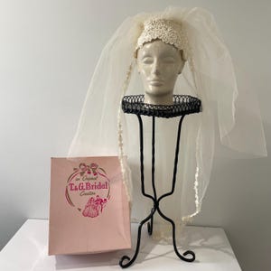 May include: A vintage bridal veil and headpiece displayed on a mannequin head atop a black metal stand. The veil is sheer, with delicate lace trim. A pink paper bag with the text "T.&G. Bridal Creation" is next to the stand.
