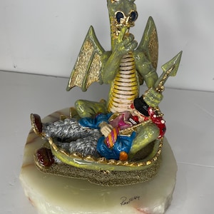 May include: A green dragon figurine with red eyes and gold-accented wings, holding a finger to its mouth in a shushing gesture. A clown figure in a blue jacket and black hat rests against the dragon. The sculpture sits on a round, pale stone base.